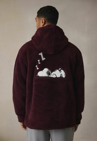 Vīna sarkanā krāsā flīsa pelēkā džemperis ar mīkstu tekstūru, uz kura izšūta Snoopy dizains, kas guļ, ar "Zzz" virs tā. Ar kapuci un vaļēju piegriezumu.
