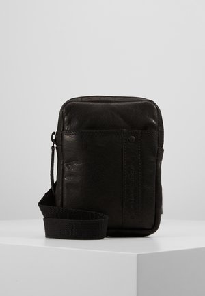 Czarna skórzana torba na ramię z przednią kieszenią, zamkiem błyskawicznym i regulowanym czarnym paskiem z materiału, stojąca na białej powierzchni na jednolitym tle.