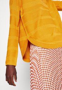 Gelber strukturierter Strickpullover mit horizontalen Streifen, kombiniert mit einem gemusterten orange-weißen Rock mit vertikalen Falten.