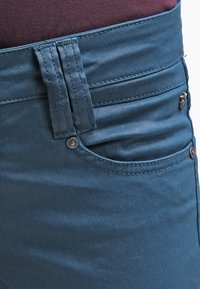 Tealblauwe skinny broek van gladde stof, met stikdetails, een voorzak en een metalen knoopaccent.