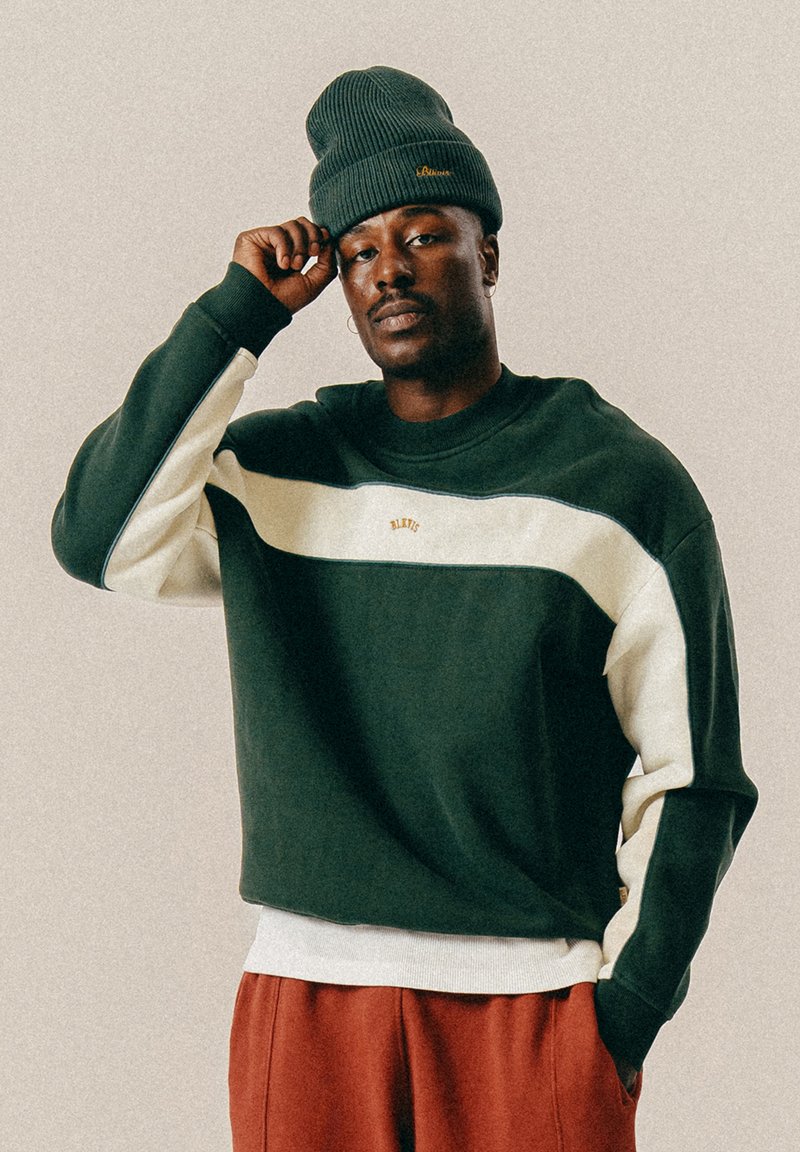 Πράσινη και κρέμα χρωματιστή μπλούζα με ρίγες και ριμπ μανσέτες και στρίφωμα; ένα υφασμάτινο σκούφο σκούρου πράσινου χρώματος με χρυσές λεπτομέρειες λογότυπου.