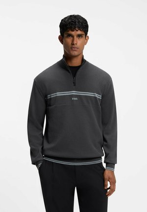 Uomo che indossa una felpa con zip grigio scuro con strisce grigio chiaro e la parola BOSS sul petto, pantaloni neri, in piedi con le mani nelle tasche.