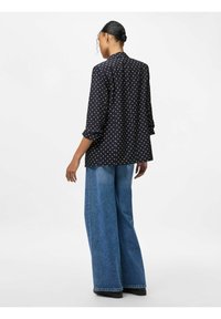 Zwarte blouse met polka dots, relaxed fit, lange mouwen opgerold en een gebogen zoom, gecombineerd met blauwe denim jeans met wijde pijpen.