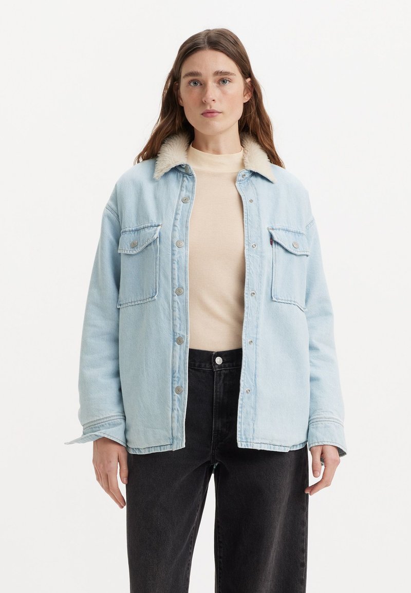 Chaqueta de mezclilla azul claro con cuello de borreguito, que presenta dos bolsillos en el pecho, cierre frontal de botones y un corte relajado, llevada sobre una blusa beige.