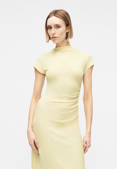 Γυναίκα με ίσια κοντά ξανθά μαλλιά που φοράει εφαρμοστό ανοιχτό κίτρινο φόρεμα με κοντά μανίκια, στέκεται μπροστά σε λευκό φόντο.