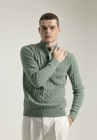 Suéter de punto trenzado en verde salvia, con cuello alto, tapeta de botones, puños y dobladillo acanalados; modelo vistiendo pantalones claros.