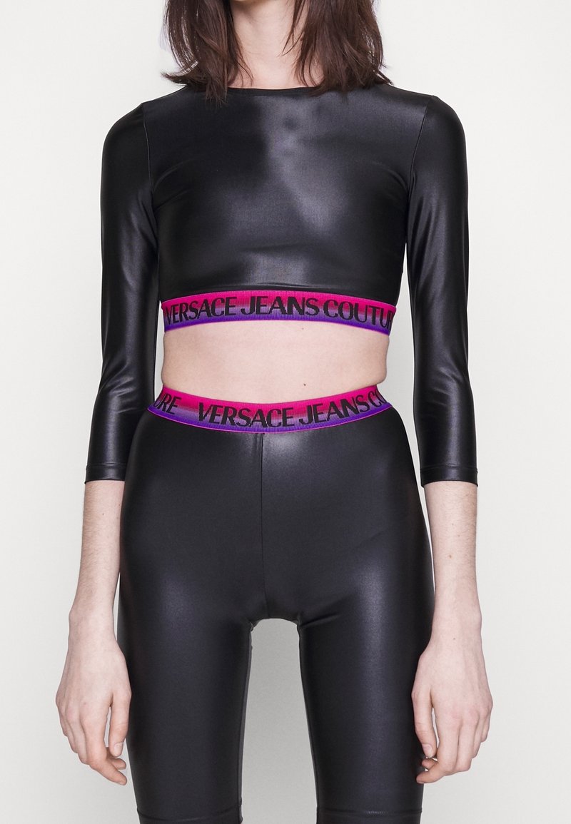 Černý bodycon top s dlouhými rukávy a zkrácenou délkou, v kombinaci s černými legínami s vysokým pasem, obě části mají růžový a fialový logový pásek.