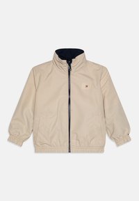 Beige Windbreaker-Jacke mit marineblauem Innenfutter, hohem Kragen, Reißverschluss, elastischen Bündchen und einem kleinen logoaufnäher auf der Brust.