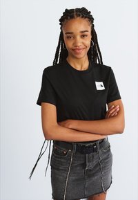 Tricou negru cu mânecă scurtă, având un patch logo alb; purtat cu o fustă din denim gri și un brâu negru, pe un fundal deschis.