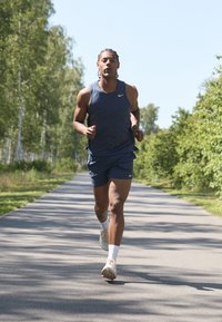 Athletic male runner in a navy sleeveless shirt and shorts, wearing light-colored sneakers, jogging on a paved path surrounded by greenery.
