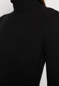 Maglione a coste nera con colletto alto e maniche lunghe, caratterizzato da un design aderente e una texture morbida. Scatto ravvicinato che mette in evidenza il colletto.