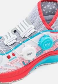 Shoe de trail running com parte superior em malha branca com detalhes em vermelho e turquesa, sistema de fecho BOA e sola texturizada para aderência.
