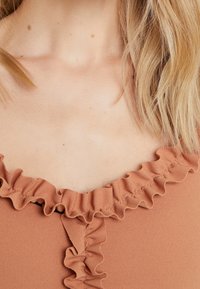 Top v rezavé barvě s volánkovým výstřihem a strukturovanou tkaninou. Detailní pohled zdůrazňuje detaily volánku a hladký povrch.