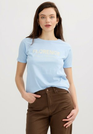 Joven con cabello castaño, viste una camiseta azul claro "Florence" y pantalones marrones, de pie con una mano en el bolsillo frente a un fondo liso.