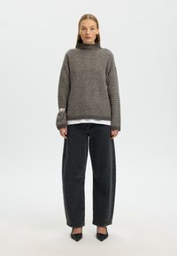 Maglione grigio a maglia con motivo a chevron e collo alto, sovrapposto a una camicia bianca, abbinato a jeans neri larghi e tacchi neri.