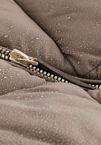 Gros plan d'une veste en tissu marron mouillé avec des gouttes d'eau et une fermeture éclair métallique partiellement ouverte.