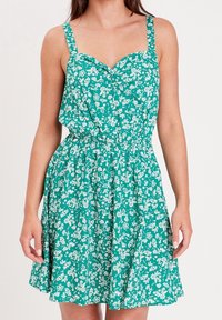 Robe florale verte avec motifs blancs, corsage ajusté, larges bretelles et taille élastique. Jupe évasée se terminant au-dessus du genou.