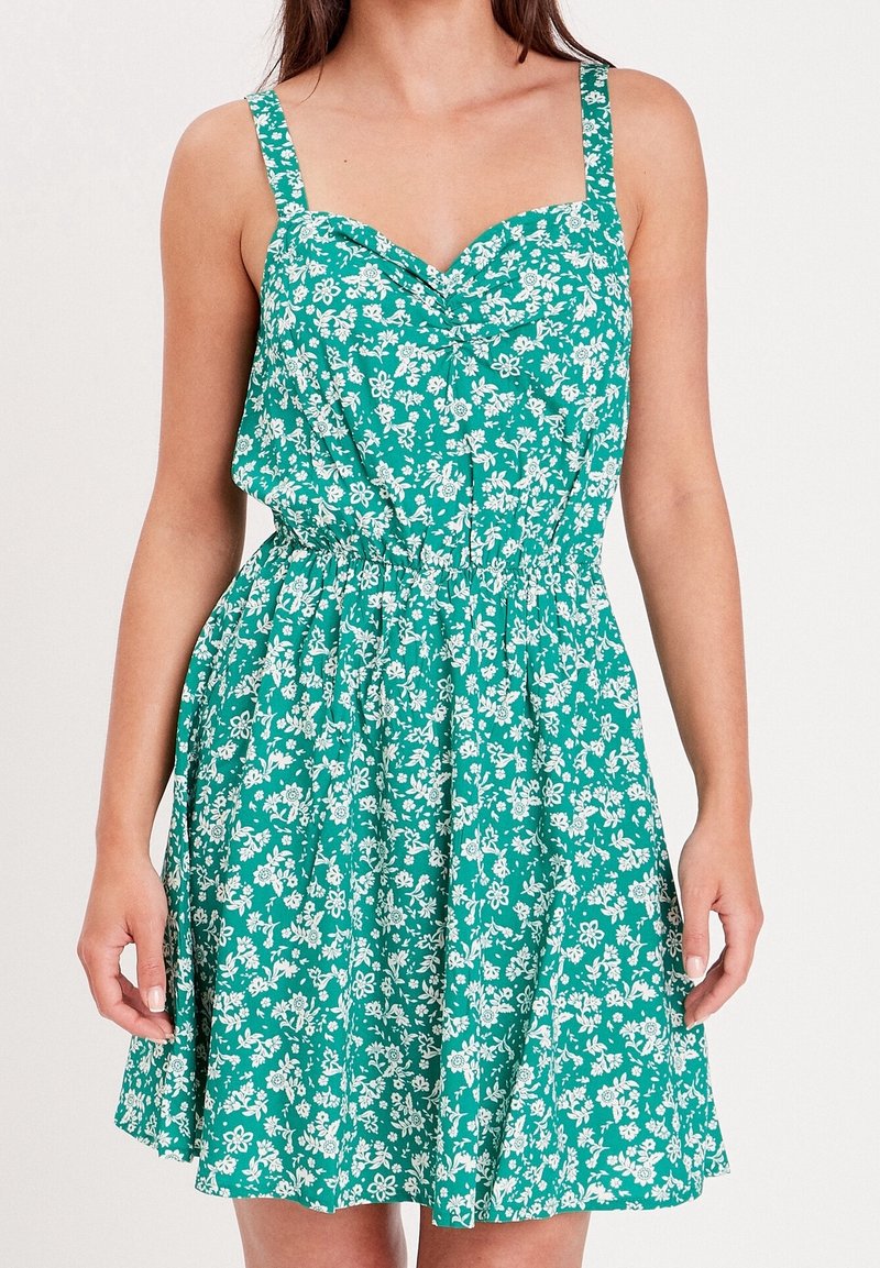 Robe florale verte avec motifs blancs, corsage ajusté, larges bretelles et taille élastique. Jupe évasée se terminant au-dessus du genou.