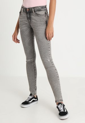 Donna con jeans skinny grigio chiaro con lievi usure sulle ginocchia e sneakers nere basse con lacci e suola bianchi.