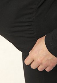 Czarna elastyczna tkanina z marszczonym wykończeniem w talii, na którym widać rękę trzymającą materiał. Design jest dopasowany i gładki.