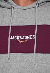 Sudadera gris con una franja burdeos en el pecho. Presenta el logo blanco "JACK&JONES" y "Originals" en naranja. Capucha con cordones ajustables.