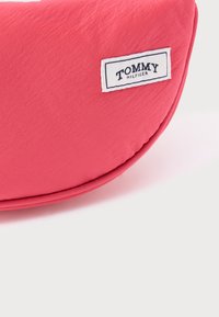 Różowa pikowana torba na ramię o teksturowanej powierzchni, z białą metką z napisem "TOMMY HILFIGER", umieszczoną na zaokrąglonym brzegu.