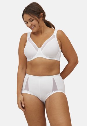 Kvinde med brunt hår samlet bagpå iført hvid støttende bh og højtaljede trusser med mesh-paneler, smilende og kiggende ned.