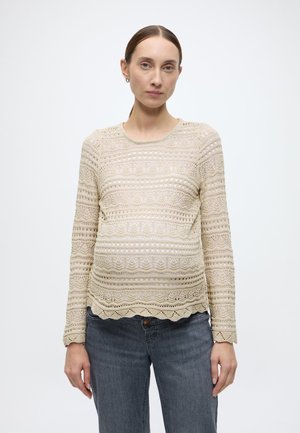 Maglione beige lavorato a maglia con motivi traforati intricati, scollo rotondo, maniche lunghe e orlo smerlato, abbinato a jeans grigi.