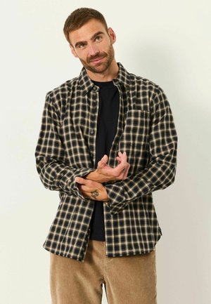 Mann mit kurzen braunen Haaren, der ein schwarz-beiges kariertes Hemd über einem schwarzen Shirt trägt, richtet seinen Ärmel und steht vor einem hellen Hintergrund.