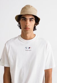 Hvid bomulds t-shirt med BMW Motorsport logo, parret med en beige bucket hat. Baggrunden er ensfarvet og neutral.