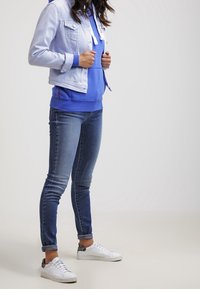 Chaqueta de denim azul claro, sudadera azul, jeans azul oscuro, zapatillas blancas con acentos grises; las características incluyen un cierre y cordones en la sudadera.