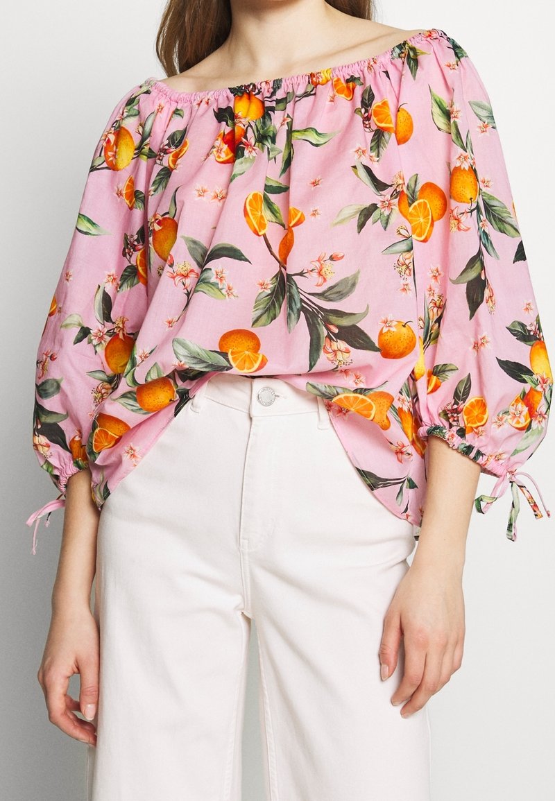 Donna che indossa una blusa rosa scollata sulle spalle con stampa di frutti arancioni e fiori, abbinata a pantaloni bianchi a vita alta.
