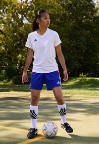 Adidas baltā īsā piedurkņu krekls, zilas šorti ar sarkanām akcentiem, melnas futbola kurpes un ceļgalus sasniedzējas baltas zeķes ar melnām svītrām, uz lauka.