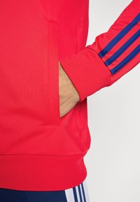 Rød sportsjakke med en tekstureret finish, der har ribbede manchetter og en sidelomme. Blå stribede detaljer langs ærmet tilføjer kontrast.