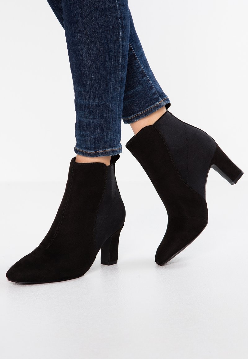 Bottines noires en mélange de daim et tissu, bout pointu, talon de 5 cm, accents sur le côté et texture lisse. Portées avec un jean bleu foncé.