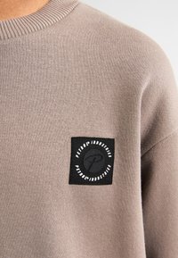 Felpa marrone con un patch rotondo in stoffa nera con testo e logo bianchi. Il materiale è lavorato a maglia, con un collo a costine.