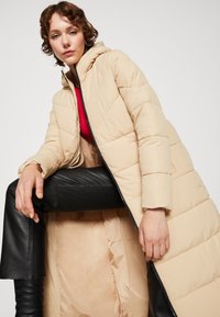 Beige dlhý puffr kabát s kapucňou, ktorý má prešívaný dizajn, predný zips a saténovú podšívku. Nosený cez čierne kožené nohavice.