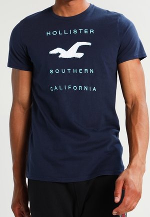 Mężczyzna w granatowym t-shircie z białym logo ptaka i turkusowym napisem "Hollister Southern California."