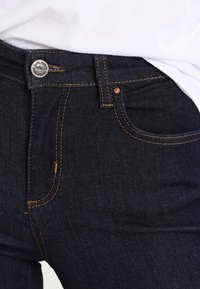 Mörkblå denimjeans med en figursydd design, med orange sömmar, en silverknapp och framfickor. Slät textur, medelvikt.