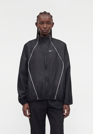 Vrouw met gevlochten haar draagt een zwarte Nike zip-up jas met witte biezen en bijpassende zwarte broek, staand tegen een effen achtergrond.