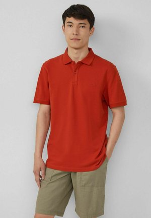 Red polo shirt with a soft texture, three-button placket, and subtle brand logo. Paired with khaki shorts featuring side pockets.