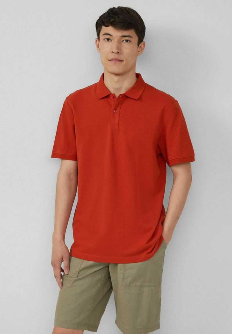 Rotes Polo-Shirt mit weicher Textur, Knopfleiste mit drei Knöpfen und subtil geprägtem Logo. Kombiniert mit khakifarbenen Shorts mit seitlichen Taschen.