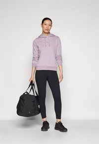 Un sweat à capuche violet clair avec logo, un legging noir et des chaussures de sport noires. Tenant un sac de sport noir, debout contre un fond neutre.