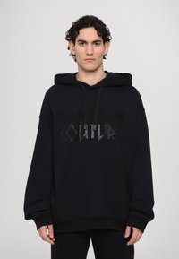 Felpa nera con un grande motivo in rilievo che recita "VERSACE KAIN COUTURE." Presenta un cappuccio con coulisse e una tasca frontale a marsupio.