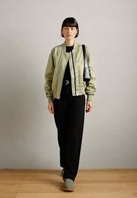 Chaqueta bomber verde oliva claro con cierre delantero, combinada con una blusa negra y pantalones de pierna ancha negros, que cuentan con un bolso negro y calzado gris.