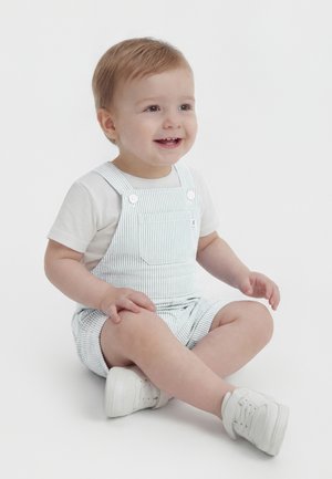 Copil mic zâmbitor cu păr castaniu deschis, purtând un tricou alb, salopetă cu dungi albastre deschise și adidași albi, stând cu picioarele încrucișate pe un podea albă.