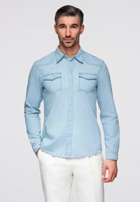 Chemise en denim bleu clair avec fermeture à boutons sur le devant, deux poches poitrine et manches longues. Présente un léger délavage et un empiècement de style western.