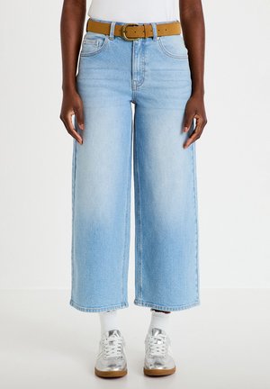 Person trägt hellblaue Jeans mit weitem Bein, einen beigefarbenen Gürtel, weiße Socken, silberne Sneaker und ein weißes Oberteil, stehend vor einem schlichten Hintergrund.