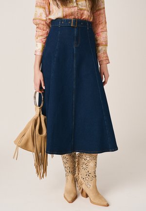 Mujer con una falda vaquera azul en línea A con cinturón, botas de ante beige con estampado, una blusa color melocotón con estampado y sosteniendo un bolso marrón con flecos.