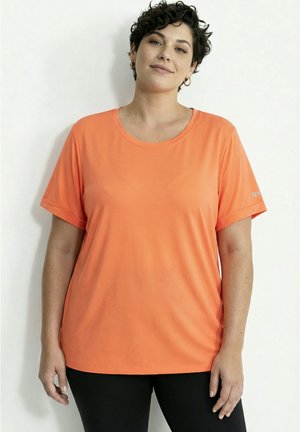 Kvinde med kort krøllet hår iført creoløreringe, en orange t-shirt med korte ærmer og sorte bukser, stående foran en hvid baggrund.